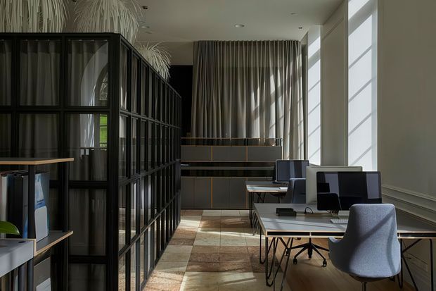 A refined, modern office interior with several workstations, black-framed glazed partition shelving, sculptural upholstered office chairs, built-in cabinetry, full-height curtains, and tall clerestory-style windows. The space has warm timber flooring, abundant natural light, and a restrained neutral palette with black accents.
