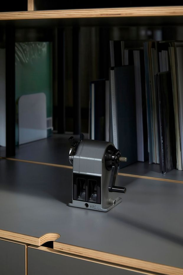 Close-up product-style photograph of a vintage manual desktop pencil sharpener placed on a dark matte built-in desk or shelf. Behind it is dark shelving with books or sample boards, while exposed layered plywood edges add a warm contrasting detail to the cabinetry. The scene is dimly lit with a refined, moody studio aesthetic.