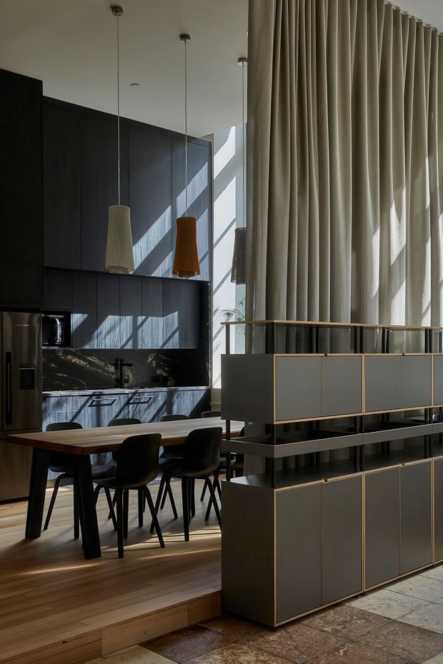 A refined open-plan kitchen and dining area with dark full-height cabinetry, a dark stone or marble-look splashback, and a timber dining table with black chairs. Three sculptural pendant lights hang above the kitchen and dining zone. A large curtain wall with tall pleated curtains brings in strong daylight, while a substantial dark built-in room divider or sideboard with warm metal trim separates the spaces. Timber flooring transitions to large-format stone or terrazzo-like flooring in the foreground.