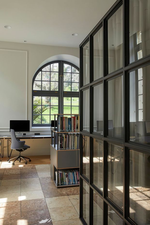 A bright, refined home office with a built-in desk beneath a large arched, gridded window, a task chair, computer monitor and freestanding bookcase. A black-framed glazed partition with multiple rectangular panes dominates the right side, separating the office from an adjacent space. The room has pale walls, warm timber flooring near the desk, large-format stone or terrazzo-like floor tiles, and abundant natural light.