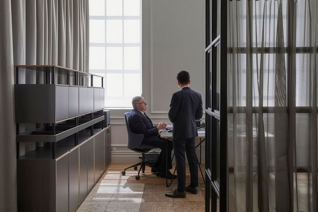 A refined office or commercial workspace with two professionals seated and standing at a desk. The room features tall windows with sheer curtains, abundant natural light, dark built-in cabinetry and shelving, polished stone or tile flooring, and glazed black-framed partitions creating a sophisticated, formal atmosphere.