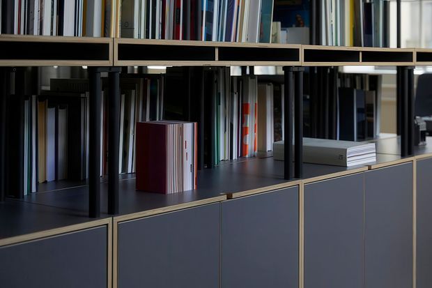 Close-up view of a sophisticated built-in shelving and storage system, with dark cabinetry, open compartments filled with books and material samples, slim brass-toned trim, and a dark countertop surface. The composition emphasizes the joinery and organized display storage.