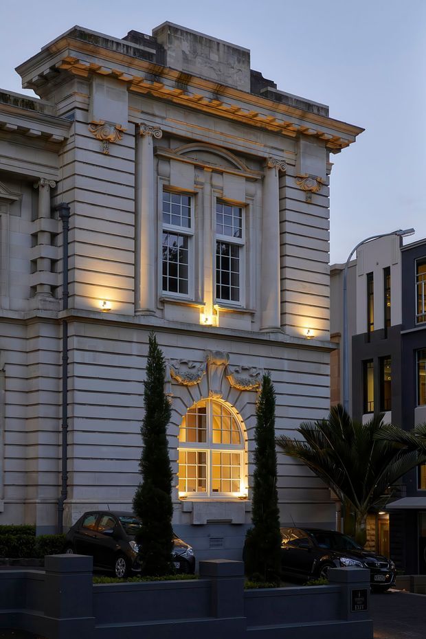 Street-level exterior view of a grand neoclassical-style building with a pale grey stone or rendered facade, strong horizontal rustication, ornate classical columns and capitals, decorative cornice, arched window surround, and tall multi-pane windows glowing with warm interior light. A contemporary neighbouring building, planted frontage, and parked cars are also visible.