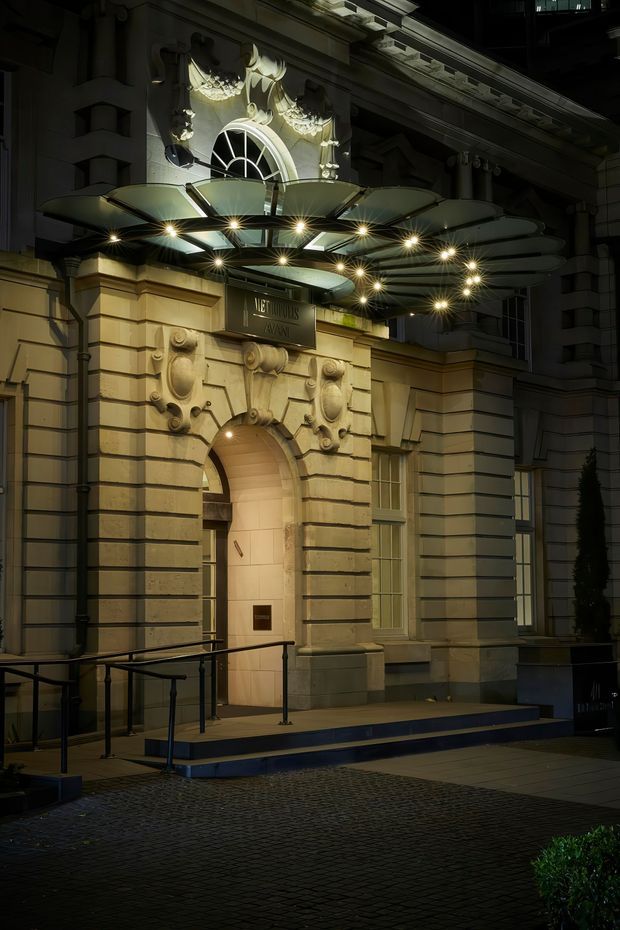 Night-time exterior view of an ornate historic commercial building entrance, featuring a richly decorated stone or rendered facade, arched doorway, tall windows, classical detailing, and a large illuminated radial canopy over the entry. Steps, accessibility ramp railings, and dark paving are visible in the foreground.