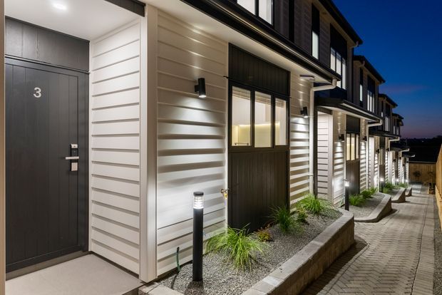 A dusk exterior view of a contemporary multi-unit residential development, showing a row of townhouse-style entrances with dark timber-look doors, white horizontal weatherboard cladding, black-framed windows, exterior wall lights, landscaped gravel beds, and a paved pedestrian walkway.