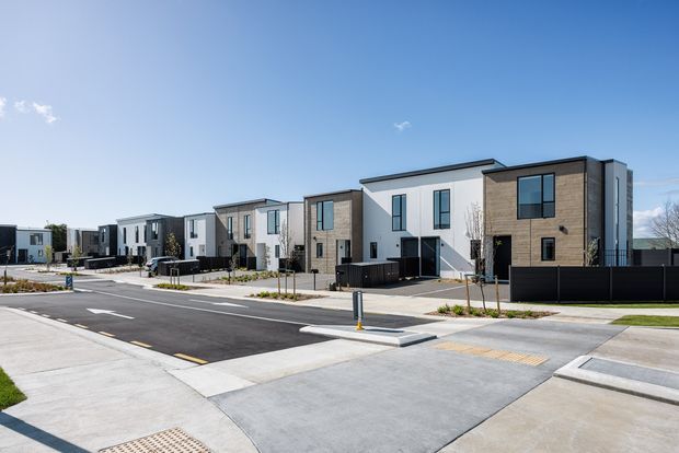 A streetscape view of a newly developed row of contemporary multi-storey townhouses. The homes feature crisp white rendered walls, dark window frames, warm timber-look facade panels, low-pitched rooflines, private fenced front courtyards, and wide concrete driveways along a newly paved residential street.