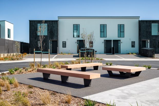 Contemporary multi-unit residential development viewed from a landscaped communal courtyard. The buildings have simple rectilinear forms, flat roofs, dark vertical cladding panels, pale rendered walls, and tall narrow windows. In the foreground are modern timber-topped benches with dark geometric supports, paved paths, low planting, and exposed aggregate or asphalt surfacing.