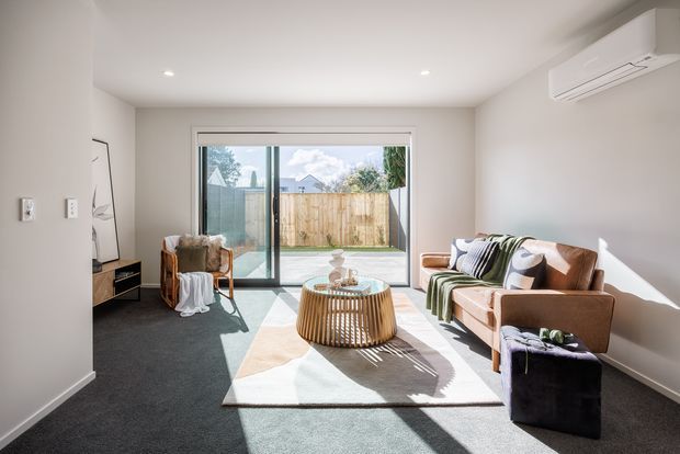 A bright, compact contemporary living room with a sofa, armchair, round timber coffee table, rug, and ottoman. Large sliding glass doors open to a small paved outdoor area, while recessed ceiling lights and a wall-mounted split-system air conditioner provide a clean modern fit-out. Neutral walls, dark carpet, warm timber, and strong natural sunlight create a relaxed, inviting atmosphere.