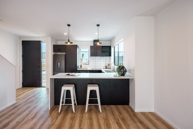 Bright contemporary open-plan kitchen viewed from the adjoining living or entry area. The kitchen features a dark wood-toned island with a light stone benchtop, matching dark cabinetry, white tiled splashback, stainless-steel appliances, pendant lighting, and warm timber flooring. A hallway and exterior doorway are visible to the left.