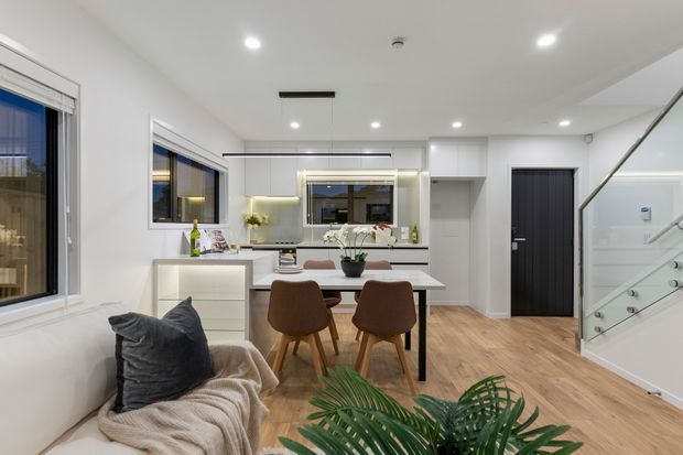 Bright open-plan residential interior combining a kitchen, dining area and living seating. The space features white cabinetry and walls, warm timber flooring, a compact dining table with upholstered chairs, a pale sofa, recessed ceiling lighting, large windows, and a contemporary staircase with glass balustrading.