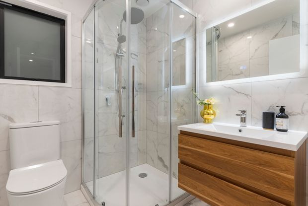 Bright contemporary bathroom featuring a frameless glass corner shower, wall-hung timber vanity with white countertop, large backlit mirror, white toilet, and white marble-look wall and floor tiles. Chrome fixtures and warm wood cabinetry add contrast to the predominantly white space.