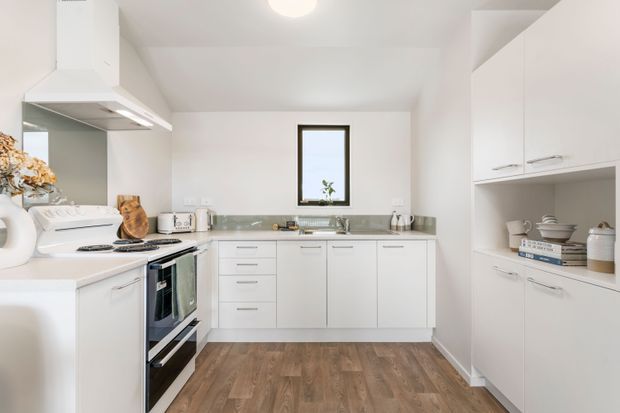 Bright, compact contemporary kitchen with white cabinetry, integrated open shelving, stainless-steel appliances, a pale tiled backsplash, and a warm wood-look floor. The U-shaped layout includes a freestanding range and sink beneath a narrow window, with simple neutral styling and abundant built-in storage.