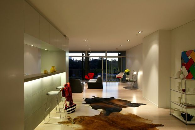 A refined contemporary open-plan interior with a minimalist kitchen and illuminated breakfast bar on the left, flowing into a spacious living area. Floor-to-ceiling glazing opens to a leafy outlook, while sculptural lounge chairs, a glass coffee table, artwork, shelving, and layered cowhide rugs add visual interest. The palette is warm and neutral with pale timber flooring, cream walls, and integrated lighting.
