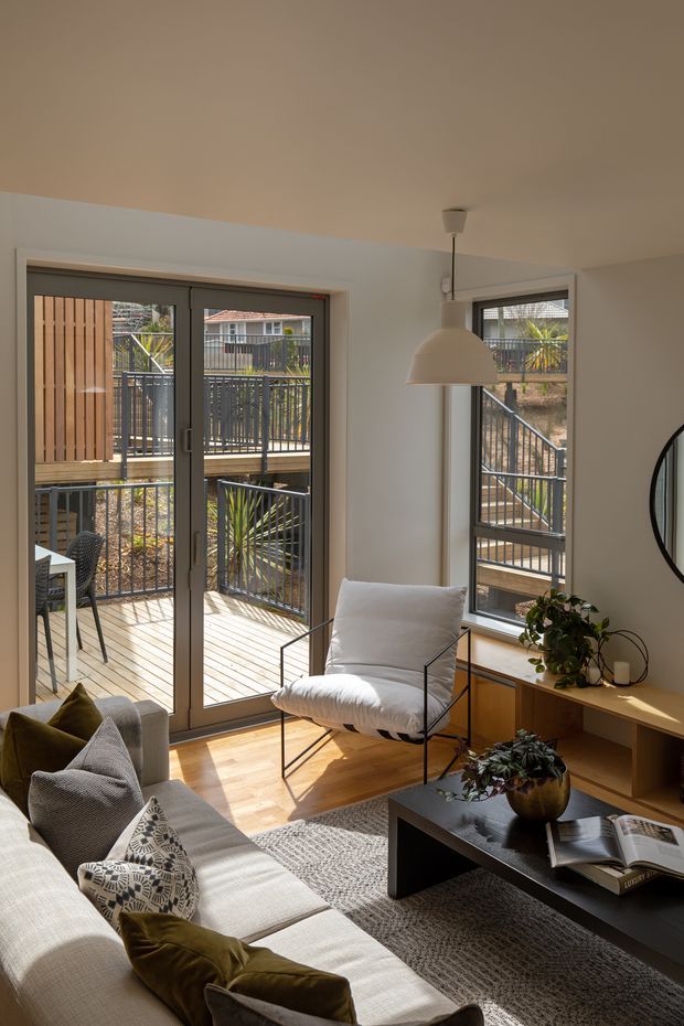 A bright, compact living room with a pale upholstered sofa, patterned and olive cushions, a black rectangular coffee table, a white lounge chair, and a low timber console. Full-height glazed doors and windows open onto a timber deck and metal-railed exterior walkway, bringing in strong natural light. The room has light timber flooring, white walls, a neutral rug, a simple pendant light, and indoor plants.
