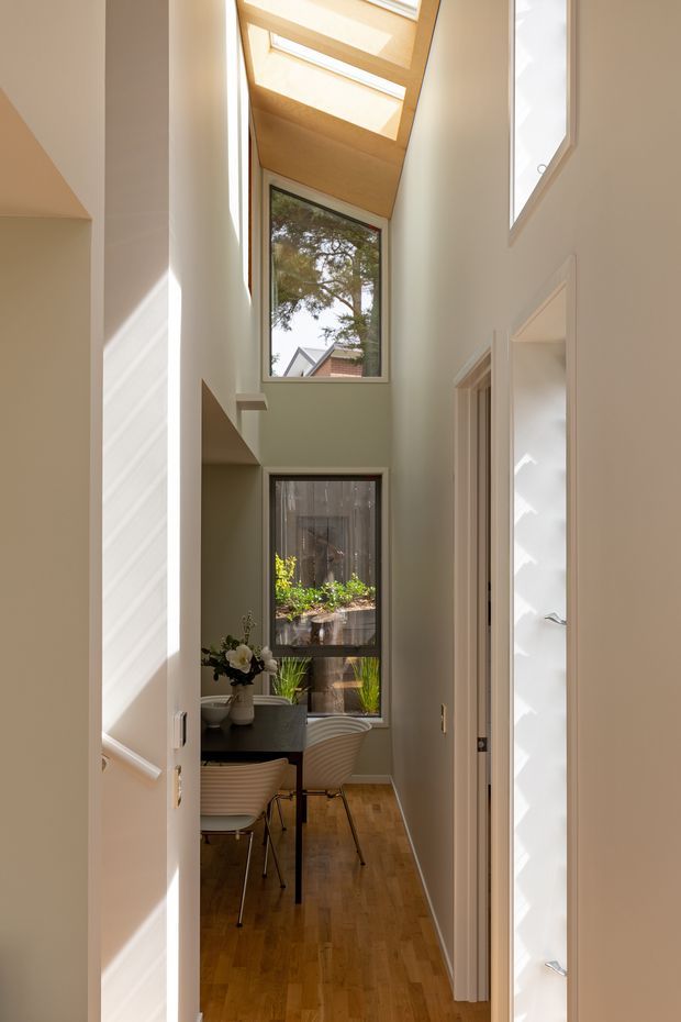 A narrow, bright interior hallway or landing overlooking a compact dining area. The space features tall white walls, a warm timber floor, high-level glazing, and a timber-lined skylight that brings in abundant natural light. A small dining table with sculptural chairs is visible at the end, along with garden views through the windows.