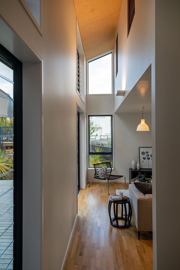 A narrow, double-height residential living area viewed from an adjoining hallway or entry. The space features warm timber flooring and a timber-lined ceiling, white walls, tall black-framed windows, a small seating area with a sculptural lounge chair and sofa, a round side table, pendant lighting, and built-in shelving. An outdoor deck and planted garden are visible through the glazing.