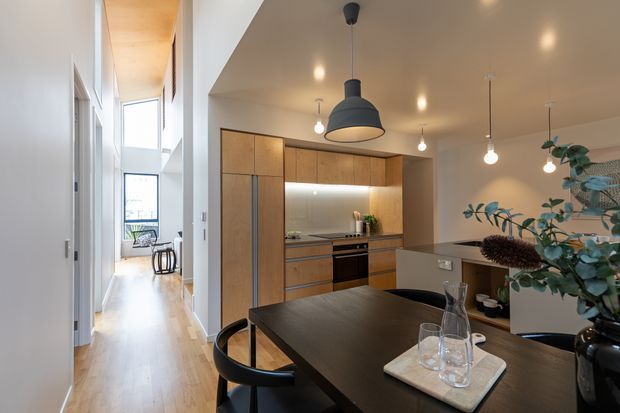 A contemporary open-plan kitchen and dining area viewed from a hallway. The kitchen features warm timber cabinetry, integrated appliances, a dark island bench, and concealed under-cabinet lighting. A dark dining table with black chairs occupies the foreground, while pendant lights and recessed ceiling lights provide illumination. A tall glazed stair or circulation area is visible to the left, with indoor greenery and tableware adding softness.