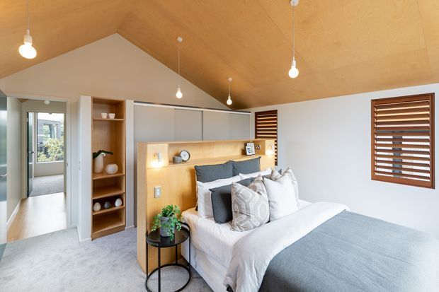 Bright, compact bedroom with a pitched plywood-lined ceiling, white walls, soft grey carpet and a neatly made bed. Built-in timber shelving and a timber headboard feature integrated bedside lighting and display niches. Warm pendant bulbs, timber venetian shutters and neutral grey-and-white bedding create a calm contemporary interior.