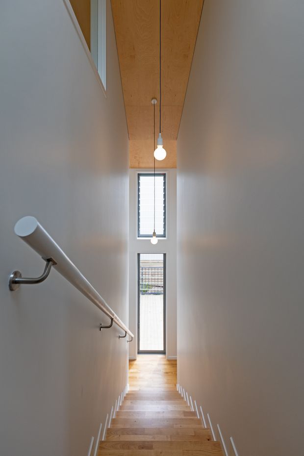 A narrow, modern stairway viewed from above, descending toward a glazed door and tall louvred window. The stair has warm timber treads, white walls, a white cylindrical handrail with metal brackets, and a warm timber-lined ceiling with simple pendant bulbs. The space is bright, streamlined, and vertically elongated.