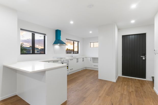 Bright contemporary open-plan kitchen and entry area with white cabinetry, a white island bench, timber-look flooring, recessed downlights, and a distinctive teal rangehood. A black vertically panelled entry door and multiple windows add contrast.