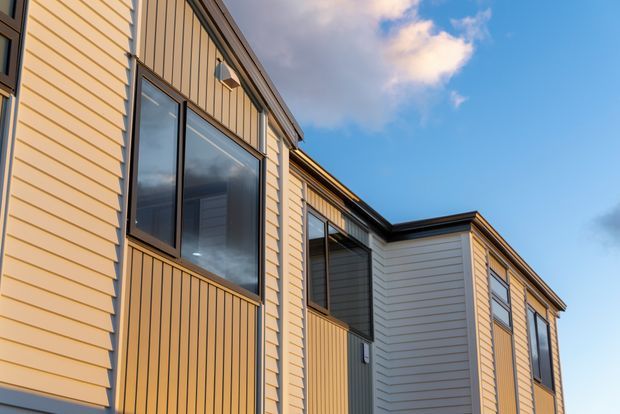 Close-up exterior view of a multi-level contemporary building with warm yellow-beige horizontal weatherboard cladding, contrasting dark vertical-panel sections, large dark-framed windows, and projecting box-like forms beneath a dark roof edge. The image focuses on the facade against a blue sky.