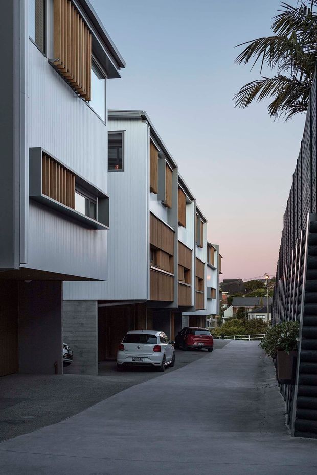 A dusk exterior view down a narrow shared driveway between contemporary multi-storey residential buildings. The left-side homes feature pale vertically ribbed cladding, projecting box forms, timber screening and balconies, with concrete garage and parking areas below. A dark repetitive facade lines the right side, while palm foliage and neighbouring houses appear in the distance.