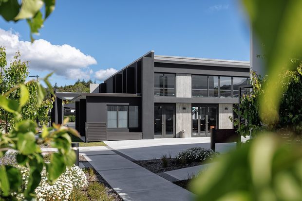 Contemporary two-storey house viewed from a landscaped front garden, with a strong geometric form, dark charcoal cladding, grey concrete-like surfaces, vertical timber-look wall panels, extensive black-framed glazing, and a flat or very low-pitched roofline. A paved path leads through the garden toward the recessed entry. The image is framed by foreground foliage under a bright blue sky.