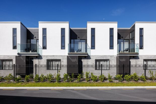 Street-facing exterior view of a contemporary multi-unit residential building with repeated white rectangular upper-level volumes, narrow vertical windows, projecting glass-balustraded balconies, dark cladding accents, and a flat roofline. A black metal fence and landscaped planting separate the building from the road.