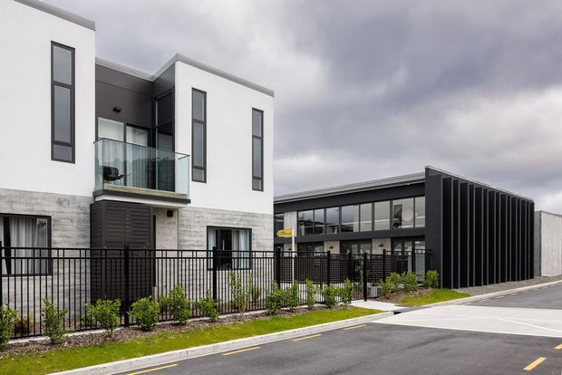 Exterior view of a contemporary multi-storey residential development beside a dark, rectilinear commercial or communal building. The white rendered facade has tall narrow windows, a glazed balcony, stone-look lower walls, and dark fencing. The adjacent building features extensive glazing and a dramatic black vertically ribbed facade. Paved roadway and landscaped planting are visible in the foreground under an overcast sky.