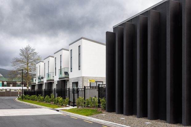Exterior view of a contemporary multi-unit residential development beside a dramatic black vertical architectural screen. The buildings have crisp white rendered facades, narrow vertical windows, projecting box forms, glass balcony balustrades, and light grey masonry at ground level. A landscaped pedestrian driveway with black metal fencing runs through the development beneath an overcast sky.