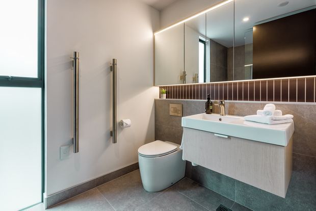 Contemporary bathroom with a wall-mounted vanity, integrated basin, wall-hung toilet, large illuminated mirror, heated towel rails, and dark grey tiled surfaces. A vertical strip of brown decorative wall tile adds contrast behind the vanity.
