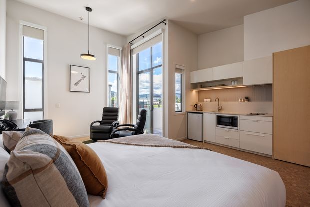 Bright, compact open-plan studio interior combining a sleeping area, lounge seating and a modern kitchenette. The room has high ceilings, large floor-to-ceiling windows and glazed doors, white walls and built-in white cabinetry with warm wood accents. A neatly made bed dominates the foreground, with two dark lounge chairs near the windows and a small streamlined kitchen along the right wall.