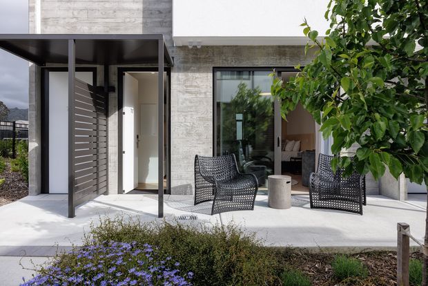 Modern residential exterior showing a covered patio or terrace beside a glazed living space. The facade combines smooth white wall planes with textured concrete or stone-like cladding, dark metal-framed glazing, and a slim flat-roof canopy. Two woven outdoor lounge chairs and a small cylindrical side table sit on a pale paved terrace, with landscaped planting in the foreground.