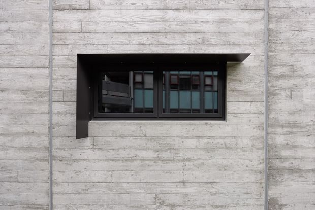 Close-up exterior view of a dark-framed horizontal window set into a light grey concrete wall. The window has a projecting black metal or dark finished surround that forms a hood and side fins, with visible glazing divided into multiple panes. The facade is minimalist and industrial in appearance, with horizontal board-like concrete texture and vertical joints.