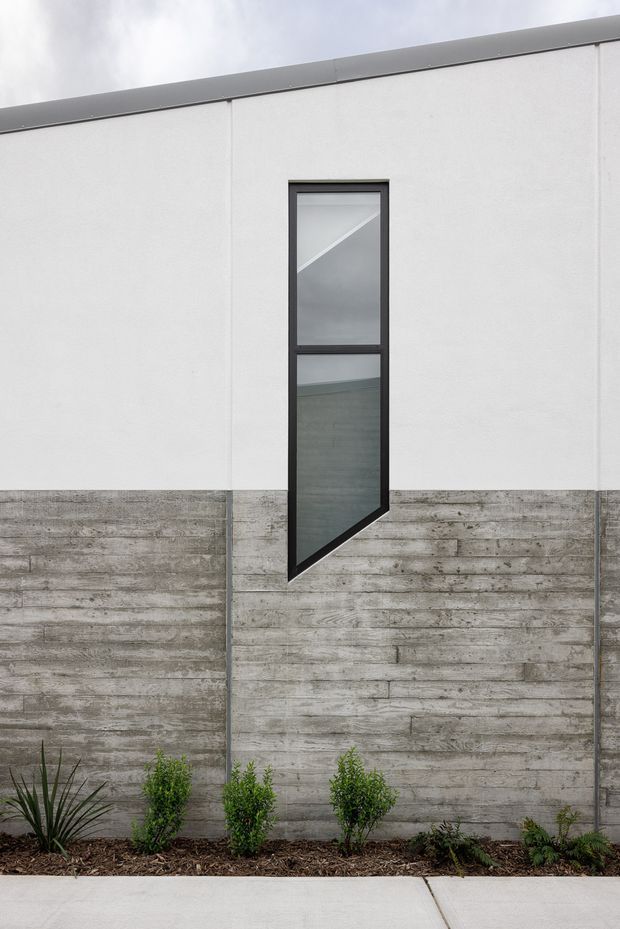 Minimalist exterior facade with a stark white rendered upper wall, rough horizontal board-formed concrete lower wall, and a tall narrow dark-framed window with an angular lower section. Low landscaping borders a concrete footpath in the foreground.