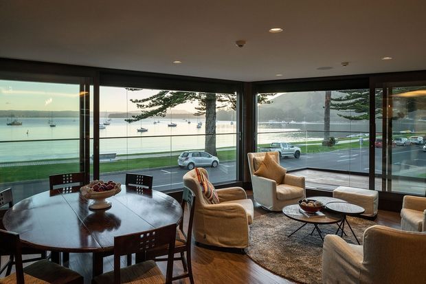 A refined living and dining room with expansive corner glazing overlooking a waterfront promenade, bay, and moored boats. The room features a dark timber dining table with matching chairs, upholstered lounge chairs and sofas, a textured area rug, low round coffee tables, timber flooring, and warm recessed lighting. The glazing creates an airy connection to the coastal setting while the furnishings give the space a comfortable, sophisticated character.