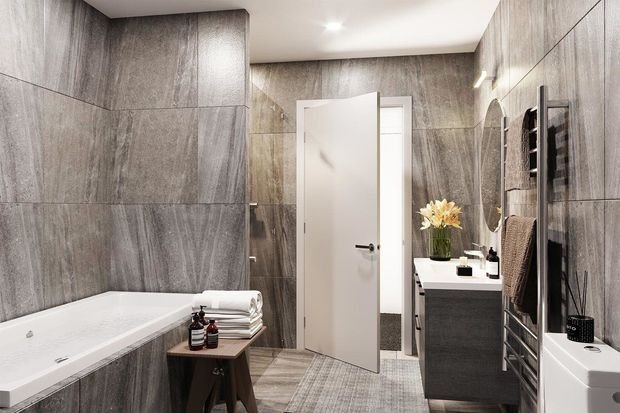 A compact contemporary bathroom with large grey stone-look wall tiles, a white built-in bathtub, grey tiled flooring, and a floating dark vanity with a white benchtop. A round wall mirror, heated towel rail, wall-mounted storage, and white toilet are visible. The room is brightly lit with a clean, refined finish.