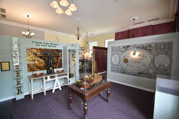 A richly detailed interior exhibition or gallery space with pale blue-grey walls, white decorative moulding, burgundy accent panels, and dark purple carpet. Display cases, framed exhibits, brass objects, and a central polished dark-wood display table fill the room. A large historic banknote-style graphic and an exhibit titled “From the River to the Bank” dominate the displays. Multiple pendant and chandelier-style lights create a warm, museum-like atmosphere.