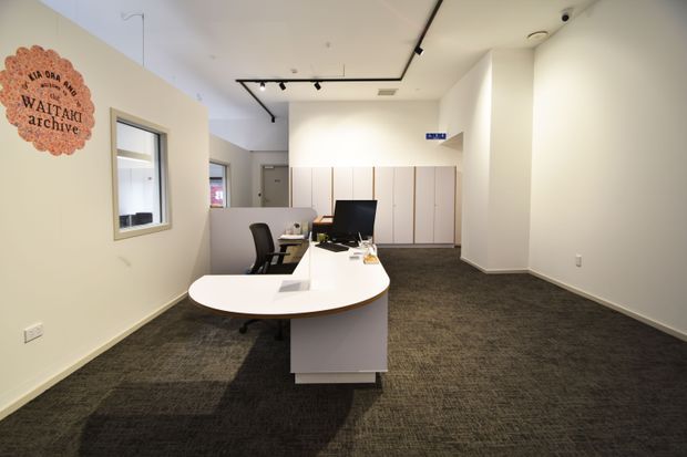 A bright, modern reception or administrative office interior with a curved white reception desk, computer workstation, visitor and staff chairs, extensive white built-in cabinetry, dark carpet flooring, and track lighting. The space has clean white walls, simple contemporary finishes, and a branded wall sign, creating a functional commercial workplace atmosphere.