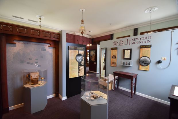 Interior exhibition or museum-style display room featuring wall-mounted historical information panels, framed documents, maps, and freestanding glass display cases. The space has deep burgundy carpet, pale blue-grey walls, dark timber trim and cabinetry, decorative pendant lights, and a small dark wood table. The room appears to present the history of New Gold Mountain.
