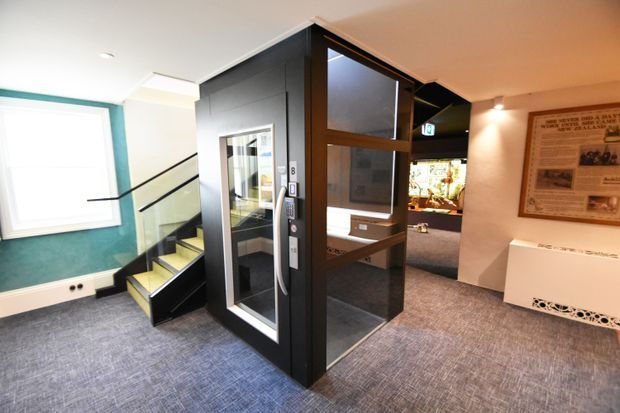 Interior view of a modern commercial or public-building entrance lobby featuring a compact glass elevator enclosed in dark timber or metal framing, adjacent stairs with pale yellow-green treads, grey carpet flooring, and a teal accent wall around a large window. A framed informational display and reception-like built-in element are visible on the right.