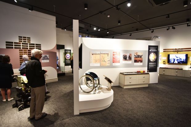 Interior view of a contemporary museum or exhibition gallery with dark ceilings, track lighting, curved white display partitions, wall-mounted informational panels, display plinths and cabinetry, and visitors viewing exhibits. The space has dark carpet flooring and a neutral white, grey, and black palette.