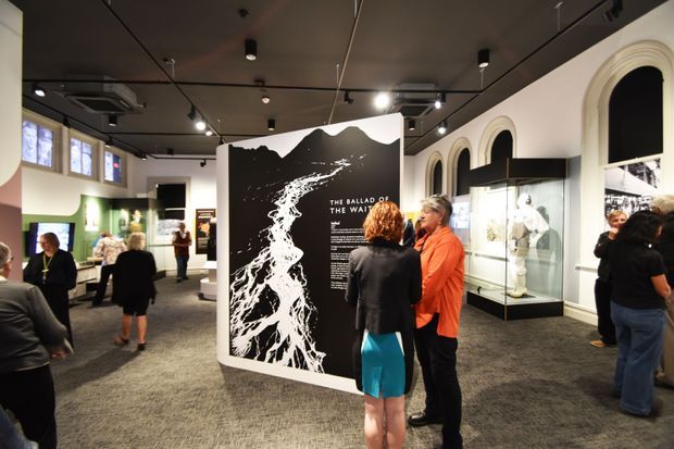 An interior exhibition or museum gallery with a dark ceiling, carpeted floor, illuminated display cases, arched wall openings, and large interpretive graphic panels. Visitors are viewing artworks or historical objects in a contemporary exhibit layout.