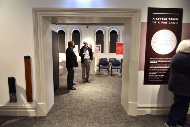 Interior view of a gallery or exhibition space framed by an ornate white moulded doorway. The room beyond has arched windows, display panels, track lighting, grey carpet, and several chairs, with visitors viewing the exhibition. A large informational wall panel is visible on the right.