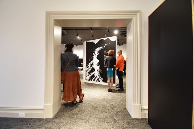 Interior view through a heavily moulded doorway into a gallery or exhibition space. White walls and layered decorative door trim frame a carpeted passage, with track lighting, large black-and-white artwork, and several visitors visible beyond. A dark partition or display wall occupies the right foreground.