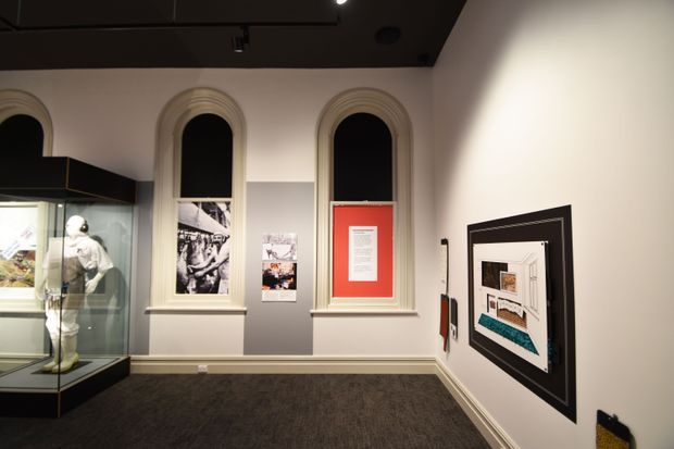 A dimly lit gallery or museum interior with three tall arched windows, ornate traditional trims, a dark ceiling, grey lower wall panels, and dark carpeted flooring. A glass display case with a mannequin stands at the left, while framed informational and architectural graphics hang on the walls.