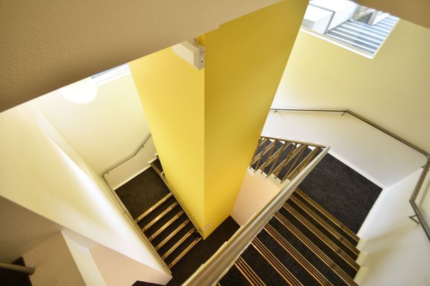 A high-angle view looking down a compact interior stairwell with multiple flights of stairs turning around a central yellow-painted structural wall. The stairs are carpeted in dark charcoal with repeated narrow yellow-gold stripes, paired with light-colored metal handrails and balustrades. White walls and ceilings surround the stairwell, with skylights bringing in natural light. The image emphasizes circulation, stair geometry, and the vertical stair void.