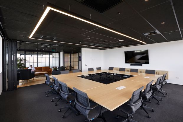 A modern commercial boardroom with a large light-wood conference table arranged around a central recessed area, surrounded by black mesh office chairs. The room has dark carpet, a black suspended ceiling with linear recessed lighting, white walls, a wall-mounted display, and glazed meeting or breakout areas beyond. A timber bench and greenery are visible in the adjoining space.