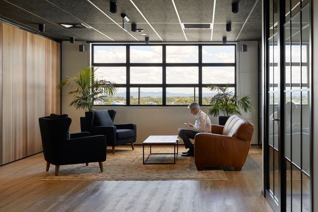 A contemporary office lounge or waiting area with several armchairs arranged around a low coffee table. The room features warm timber wall panelling, a large black-framed grid window with expansive views, exposed dark ceiling panels with track-style lighting, timber flooring, indoor palms, and a glazed black-framed partition on the right. A person is seated in one of the chairs.