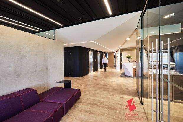 A contemporary commercial interior showing a spacious reception and circulation area. The room features warm timber-look flooring, a large textured pale wall, dark ribbed ceiling panels with linear integrated lighting, extensive glass partitions and doors, black built-in joinery, and a geometric white reception desk. A deep purple sectional sofa occupies the foreground, while workspaces are visible beyond the glazing.